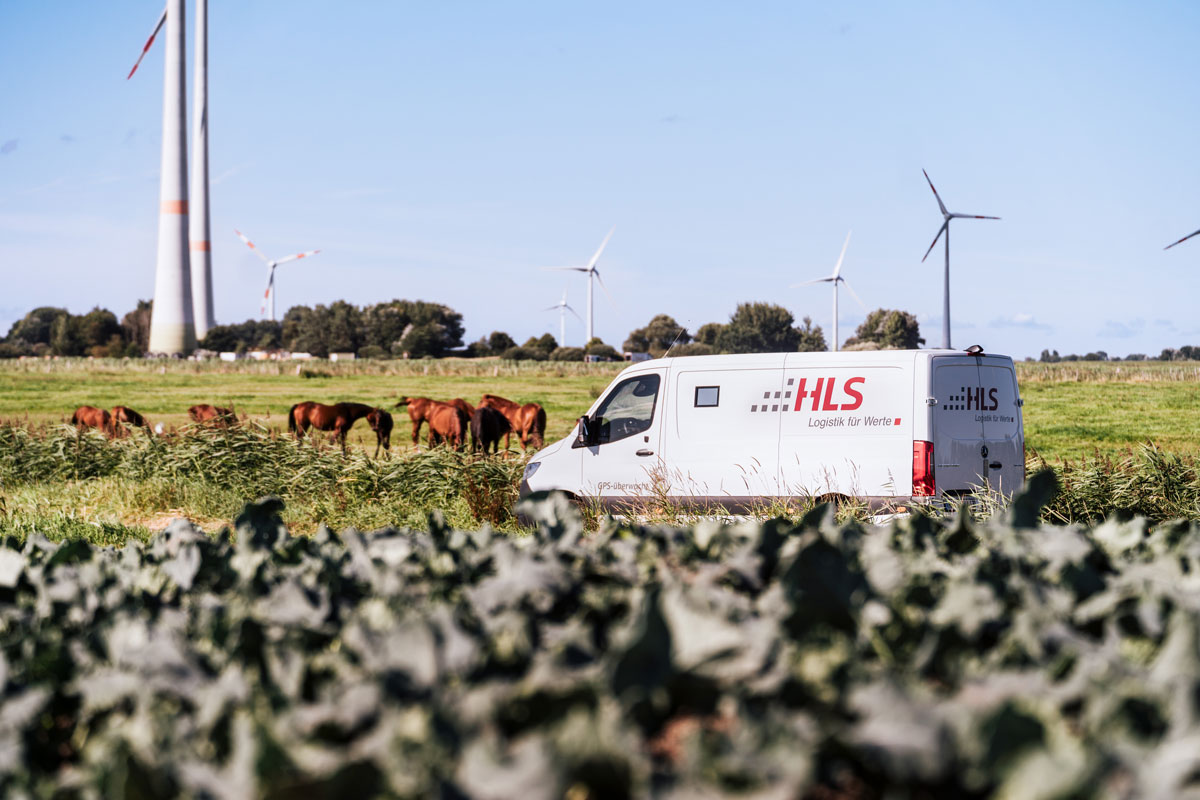 HLS-Transporter fährt durch norddeutsche Landschaft mit Windrädern und Pferden